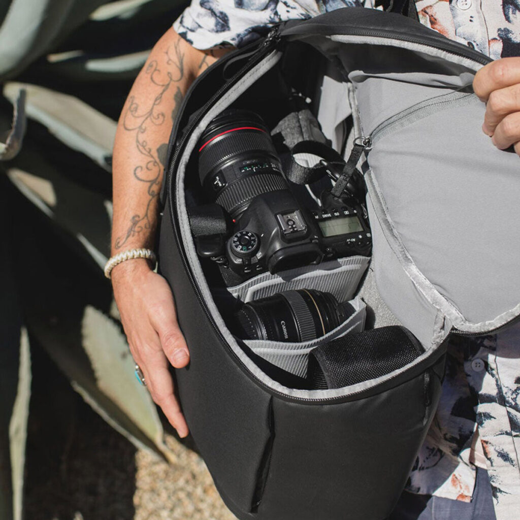 Peak-Design-Everyday-Backpack-v2-30l-black-10 Kukkaiseen paitaan pukeutunut henkilö, jolla on tatuoitu käsivarsi, pitää mustaa Peak Design Everyday Backpack v2 30L -reppua auki ja näyttää DSLR-kameran ja useita objektiiveja siististi järjestettynä sen pehmustetuissa lokeroissa.