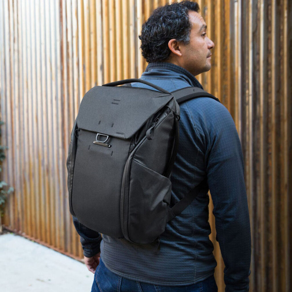 Peak-Design-Everyday-Backpack-v2-30l-black-11 Tummiin takkeihin pukeutunut mies, jolla on mukanaan Peak Design Everyday Backpack v2 30L -kamerareppu, musta, seisoo ulkona pystysuoran puuaidan vieressä ja katsoo oikealle.