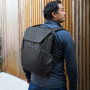 Peak-Design-Everyday-Backpack-v2-30l-black-11 Tummiin takkeihin pukeutunut mies, jolla on mukanaan Peak Design Everyday Backpack v2 30L -kamerareppu, musta, seisoo ulkona pystysuoran puuaidan vieressä ja katsoo oikealle.