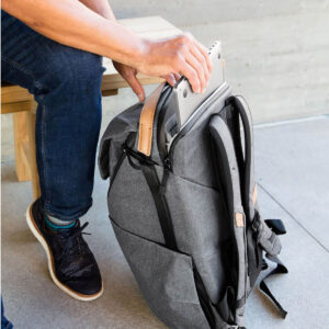 Peak-Design-Everyday-Backpack-v2-30l-charcoal-7 Farkkuihin ja mustiin lenkkareihin pukeutunut henkilö istuu penkillä ja laittaa kannettavan tietokoneen tummanharmaa Peak Design Everyday Backpack v2 30L -kamerareppu lepää maassa vieressä.