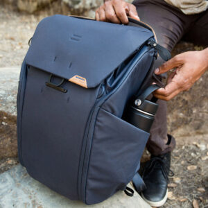 Peak-Design-Everyday-Backpack-v2-30l-midnight-8 Ulkoileva henkilö laittaa mustan vesipullon kalliolla istuvan Peak Design Everyday Backpack v2 30L -kamerareppu, tummansininen, sivutaskuun. Henkilöllä on yllään ruskeat housut ja mustat kengät.