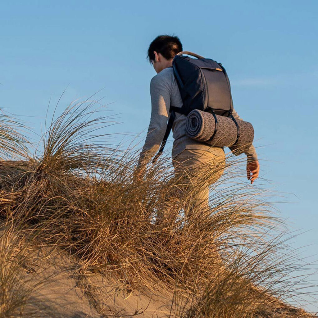 Peak-Design-Everyday-Backpack-v2-30l-midnight-9 Henkilö, jolla on mukanaan Peak Design Everyday Backpack v2 30L -kamerareppu, tummansininen ja rullamatto, kiipeää hiekkamäkeä, jossa on kuivaa ruohoa kirkkaansinisen taivaan alla.