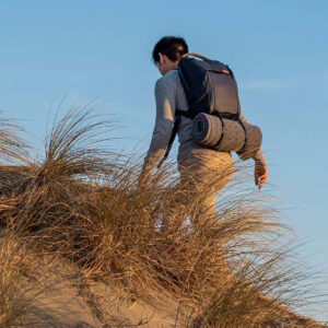 Peak-Design-Everyday-Backpack-v2-30l-midnight-9 Henkilö, jolla on mukanaan Peak Design Everyday Backpack v2 30L -kamerareppu, tummansininen ja rullamatto, kiipeää hiekkamäkeä, jossa on kuivaa ruohoa kirkkaansinisen taivaan alla.