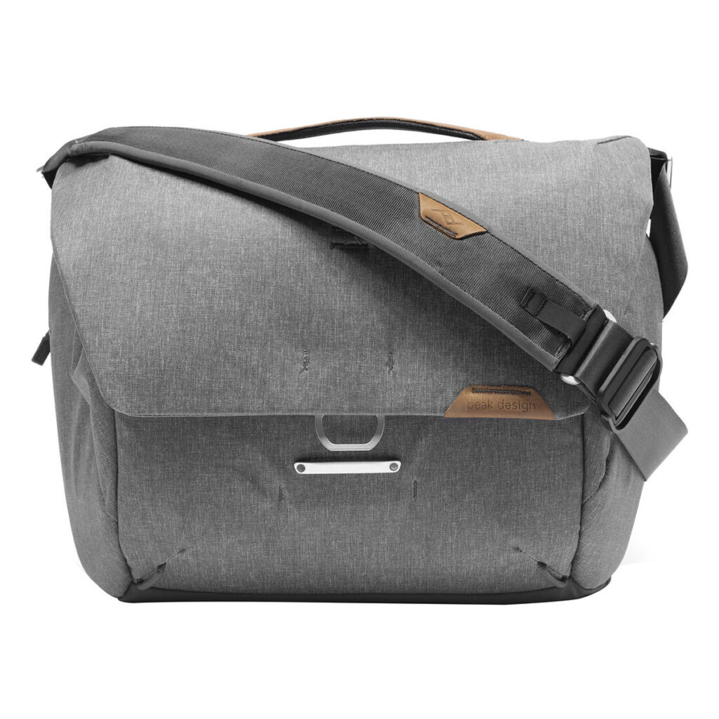 Peak-Design-Everyday-Messenger-13L-v2-Ash-2 Peak Design Everyday Messenger 13L V2 - Ash on harmaa laukku, jossa on suuri läppä, säädettävä olkahihna ja yläkahva. Laukussa on metalli- ja nahkakoristeita, ja se on ihanteellinen kannettavien tietokoneiden tai kameravarusteiden kuljettamiseen.