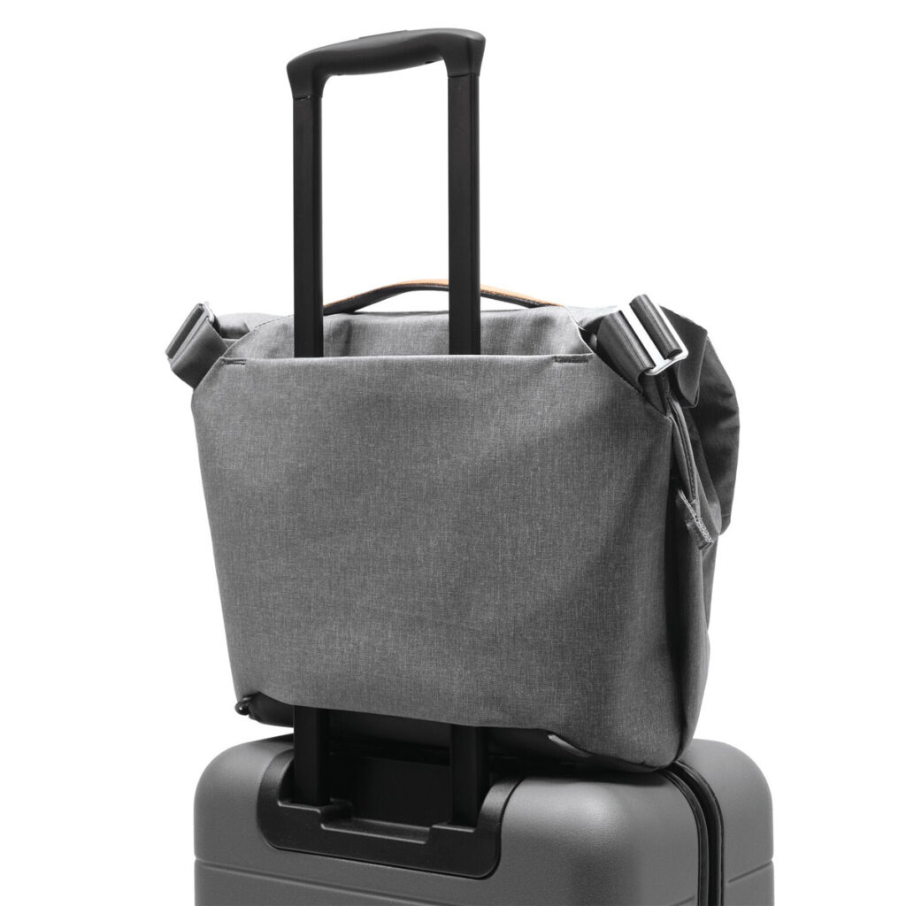 Peak-Design-Everyday-Messenger-13L-v2-Ash-6 Peak Design Everyday Messenger 13L V2 - Ash on sijoitettu turvallisesti mustan rullamatkalaukun kahvan päälle ja esittelee kätevän matkavaraston.