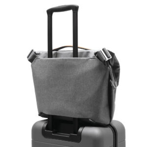 Peak-Design-Everyday-Messenger-13L-v2-Ash-6 Peak Design Everyday Messenger 13L V2 - Ash on sijoitettu turvallisesti mustan rullamatkalaukun kahvan päälle ja esittelee kätevän matkavaraston.