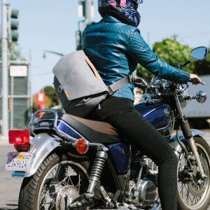Peak-Design-Everyday-Messenger-13L-v2-Ash-8 Sinisiin varusteisiin ja kypärään pukeutunut ratsastaja kantaa Peak Design Everyday Messenger 13L V2 - Ash -matkatavaraa ajaessaan sinisellä moottoripyörällä risteyksen läpi liikennevalojen ja taustalla näkyvien puiden kanssa.