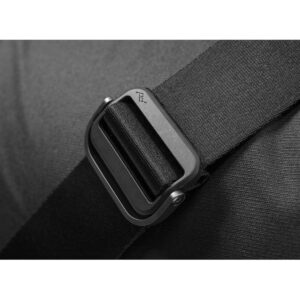 Peak-Design-Everyday-Messenger-13L-v2---Black-4 Lähikuva mustasta hihnasta, jossa on PD-logolla kaiverrettu metallisolki, kiinnitettynä Peak Design Everyday Messenger 13L V2 - Black -laukun tummaan kangaspintaan.