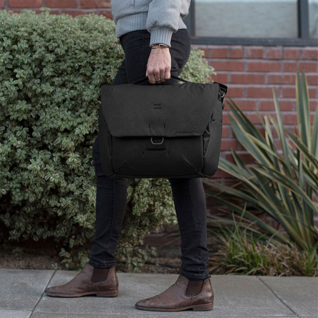 Peak-Design-Everyday-Messenger-13L-v2---Black-7 Tummiin housuihin, ruskeisiin saappaisiin ja vaaleanharmaaseen villapaitaan pukeutunut henkilö kävelee jalkakäytävällä pitäen kädessään Peak Design Everyday Messenger 13L V2 - Black. Taustalla näkyy vihreitä pensaita ja tiiliseinä.