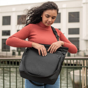 Peak-Design-Everyday-Tote-15l-black-8 Nainen seisoo ulkona kaiteen vieressä ja katsoo alaspäin, kun hän kurottautuu Peak Design Everyday Tote 15L v2 mustaan. Hänellä on punainen villapaita ja farkut. Taustalla näkyy valkoinen rakennus, jossa on paljon ikkunoita.