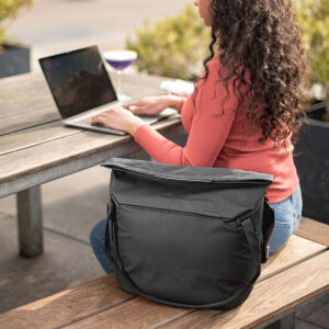 Peak-Design-Everyday-Tote-15l-black-9 Nainen, jolla on pitkät kiharat hiukset ja korallivärinen villapaita, käyttää kannettavaa tietokonetta ulkona olevan puupöydän ääressä. Peak Design Everyday Tote 15L v2 - Black lepää hänen vieressään penkillä, ja pöydälle on asetettu juoma.
