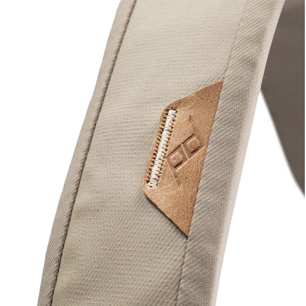 Peak-Design-Everyday-Tote-15l-bone-3 Lähikuva Peak Design Everyday Tote 15L v2 - Bone -laukusta, jossa näkyy beige kangashihna, johon on ommeltu ruskea nahkalappu ja kohokuvioitu geometrinen logo.