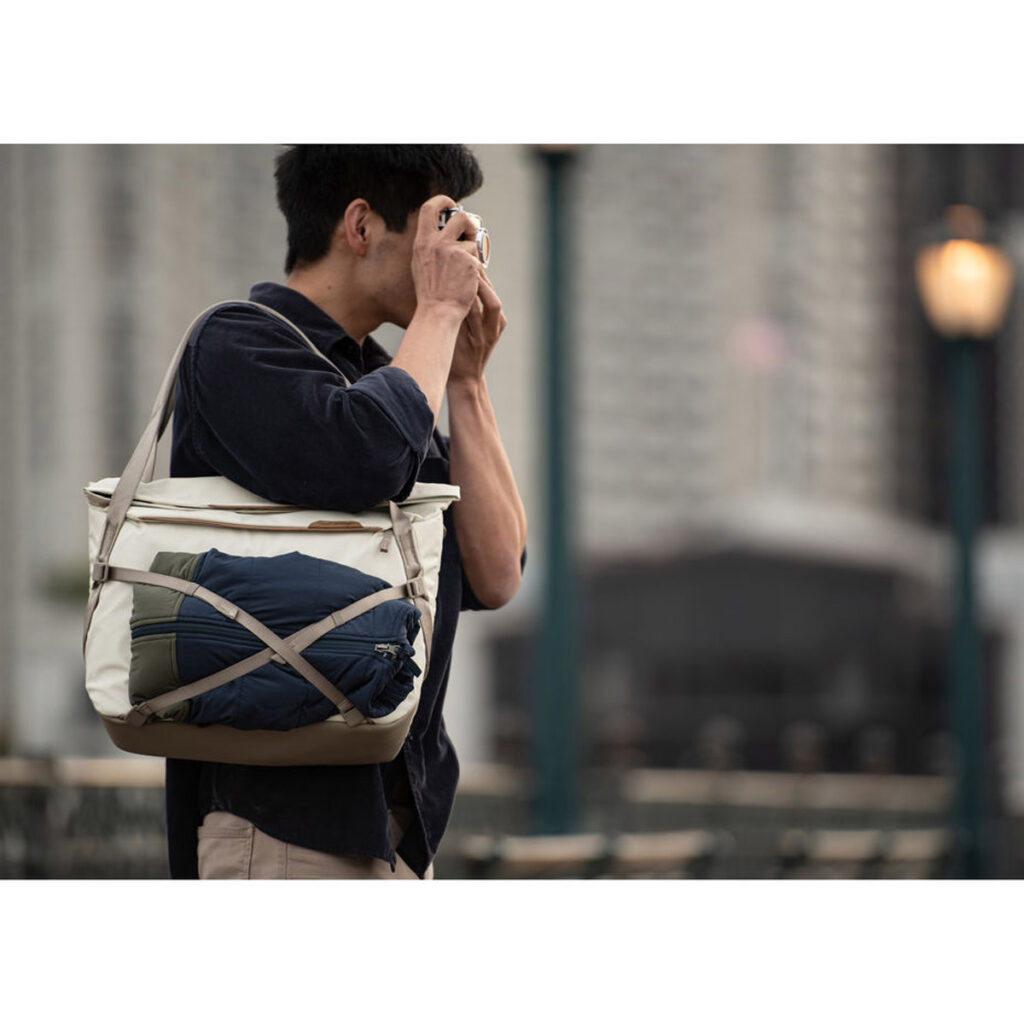 Peak-Design-Everyday-Tote-15l-bone-6 Henkilö seisoo ulkona kaupunkiympäristössä mustaan paitaan pukeutuneena, ja hänellä on mukanaan Peak Design Everyday Tote 15L v2 - Bone -rinkka, johon on kiinnitetty kääritty takki tai huopa. Taustalla näkyy rakennuksia ja lyhtypylväitä.