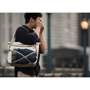 Peak-Design-Everyday-Tote-15l-bone-6 Henkilö seisoo ulkona kaupunkiympäristössä mustaan paitaan pukeutuneena, ja hänellä on mukanaan Peak Design Everyday Tote 15L v2 - Bone -rinkka, johon on kiinnitetty kääritty takki tai huopa. Taustalla näkyy rakennuksia ja lyhtypylväitä.