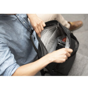 Peak-Design-Everyday-Totepack-20l-black-6 Vaaleansiniseen paitaan pukeutunut henkilö avaa mustan Peak Design Everyday Totepack 20L v2 -repun, jonka sisältä paljastuu puhelin, kamera ja omena harmaalla alustalla istuen. Vain ylävartalo ja kädet ovat näkyvissä.