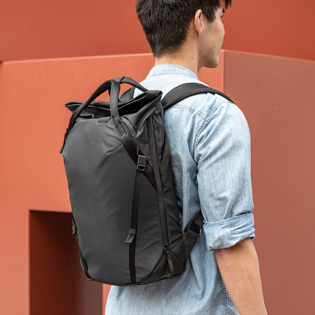 Peak-Design-Everyday-Totepack-20l-black-8 Henkilö, jolla on vaaleansininen farkkupaita ja mukanaan Peak Design Everyday Totepack 20L v2 - Black, seisoo modernin, ruosteenvärisen seinän edessä kamerasta poispäin.