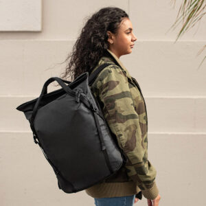 Peak-Design-Everyday-Totepack-20l-black-9 Kiharatukkainen nainen naamiotakissa seisoo sivuttain vaaleaa seinää vasten, selässään musta Peak Design Everyday Totepack 20L v2.
