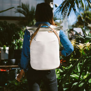 Peak-Design-Everyday-Totepack-20l-bone-8 Farkkupaitainen ja mustiin housuihin pukeutunut henkilö seisoo vihreiden kasvien keskellä, kasvot poispäin, ja kantaa Peak Design Everyday Totepack 20L v2 - Bone -reppua selässään, kun auringonvalo suodattuu lehvistön läpi.