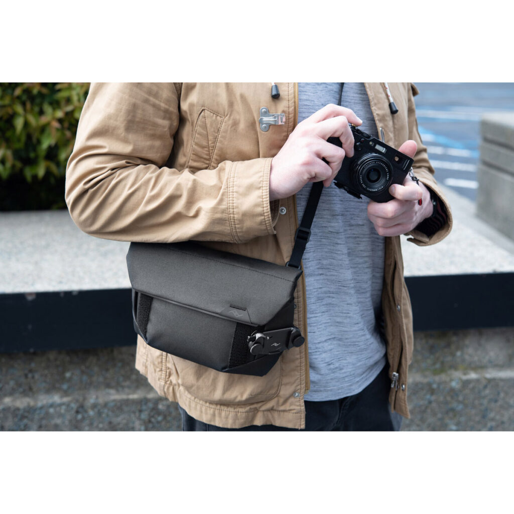 Peak-Design-Field-Pouch-V2-black-8 Ruskeaan takkiin ja harmaaseen paitaan pukeutunut henkilö pitelee kameraa, ja hänellä on vartalonsa poikki Peak Design Field Pouch V2 - Black. Taustalla näkyy betonipenkki ja viherkasveja.
