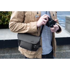 Peak-Design-Field-Pouch-V2-black-8 Ruskeaan takkiin ja harmaaseen paitaan pukeutunut henkilö pitelee kameraa, ja hänellä on vartalonsa poikki Peak Design Field Pouch V2 - Black. Taustalla näkyy betonipenkki ja viherkasveja.