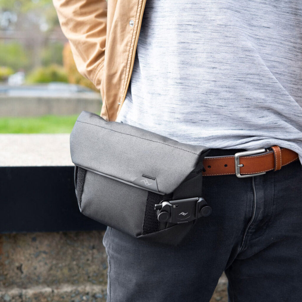 Peak-Design-Field-Pouch-V2-black-9 Ulkona oleva henkilö pukeutuu vaaleanharmaaseen paitaan, ruskehtavaan takkiin ja tummiin farkkuihin, joissa on ruskea vyö, ja hänellä on Peak Design Field Pouch V2 - Black vyötäröllä. Tausta on sumea.