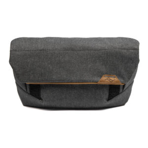 Peak-Design-Field-Pouch-V2-charcoal-1 Peak Design Field Pouch V2 - Charcoal on messenger-tyylinen laukku hiilenharmaasta kankaasta, jossa on mustat hihnat, läppäsuljenta ja pieni ruskea nahkalappu edessä, jossa on minimalistinen geometrinen logo.