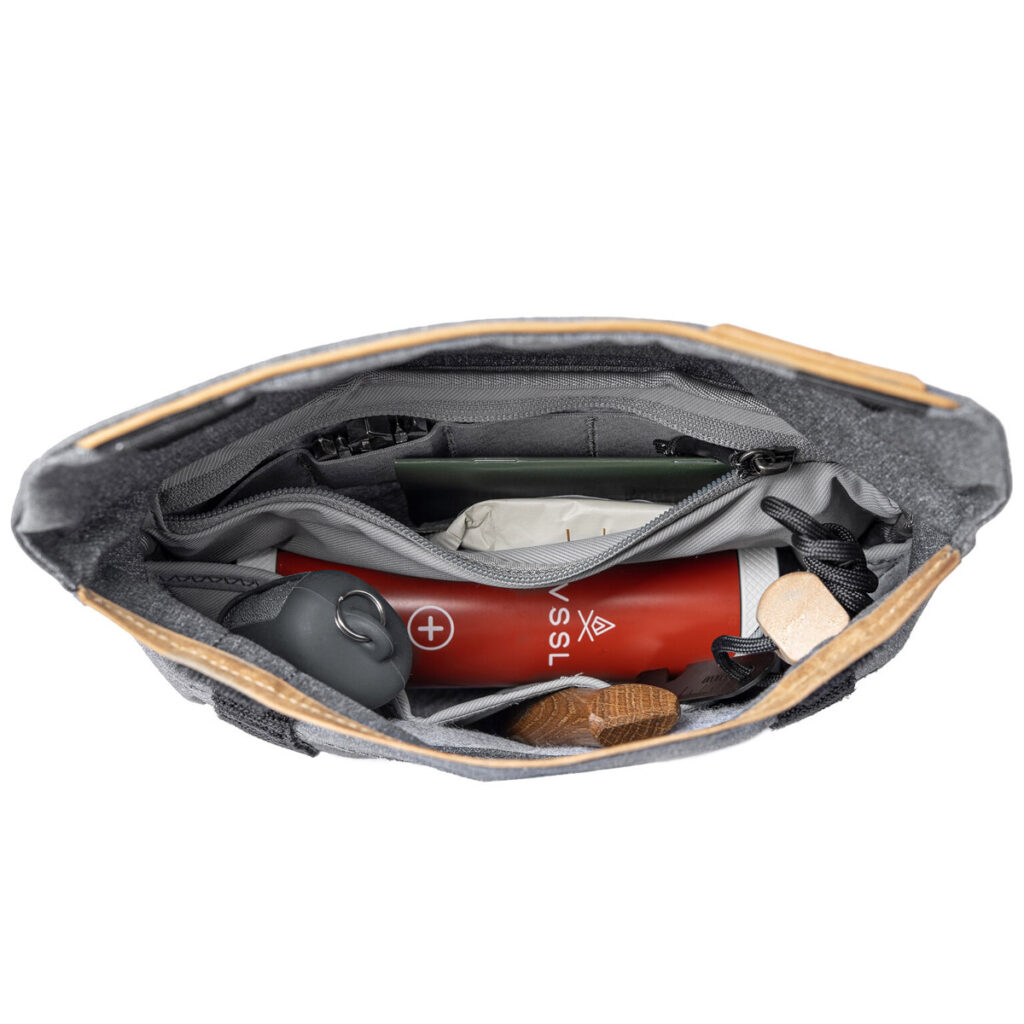 Peak-Design-Field-Pouch-V2-charcoal-4 Ylhäältä alaspäin näkymä Peak Design Field Pouch V2 - Charcoal -laukusta, joka on avattu paljastaen siististi järjestetyn sisällön, kuten punaisen ensiapupakkauksen, harmaan esineen, jossa on karabiini, puisen esineen ja useita pieniä esineitä useissa lokeroissa.