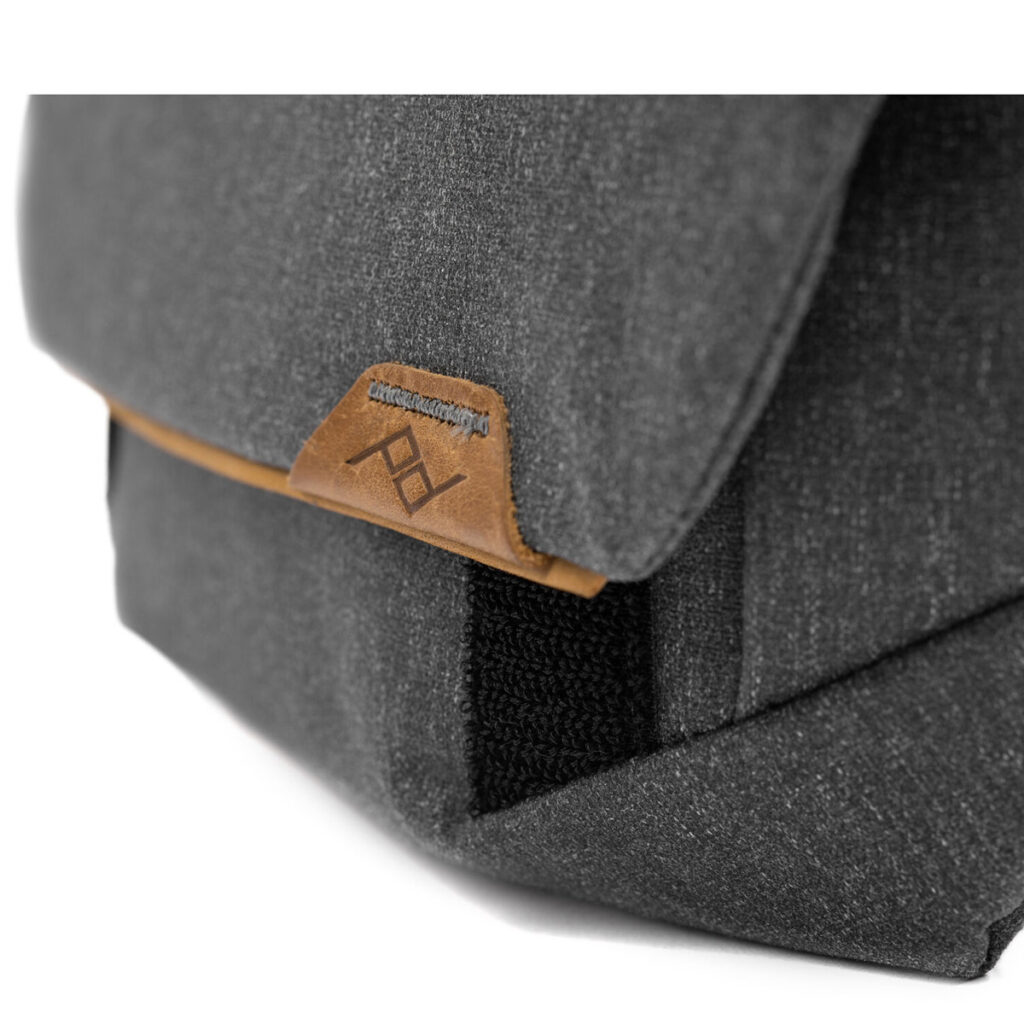 Peak-Design-Field-Pouch-V2-charcoal-5 Lähikuva Peak Design Field Pouch V2 -laukusta Charcoal, jossa näkyy sen tummanharmaa kangas ja ruskea nahkakoristeinen läppä, jossa on ommeltu logo. Materiaalin rakenne näkyy selvästi.