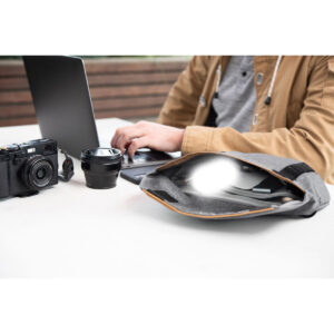 Peak-Design-Field-Pouch-V2-charcoal-8 Ruskeaan takkiin pukeutunut henkilö käyttää kannettavaa tietokonetta ulkopöydässä. Lähellä on kamera, objektiivi ja avoin Peak Design Field Pouch V2 - Charcoal, jossa on puhelin ja pientä elektroniikkaa.
