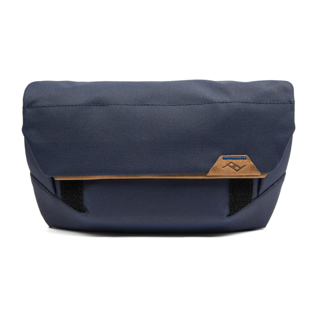 Peak-Design-Field-Pouch-V2-midnight-1 Peak Design Field Pouch V2 - Midnight on tummansininen rintareppu, jossa on etuläppä, ruskehtava nahkalappu, jossa on geometrinen logo, ja kaksi näkyvää mustaa hihnaa alareunassa.