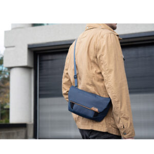 Peak-Design-Field-Pouch-V2-midnight-8 Ruskehtavaan takkiin pukeutunut henkilö seisoo rakennuksen ulkopuolella, ja hänellä on mukanaan Peak Design Field Pouch V2 - Midnight, tummansininen crossbody-laukku, jonka etupuolella on pieni ruskea logo.