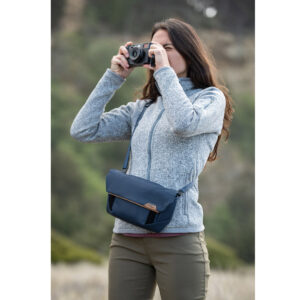 Peak-Design-Field-Pouch-V2-midnight-9 Pitkänruskeatukkainen nainen, jolla on harmaa vetoketjullinen takki ja vihreät housut, seisoo ulkona kamera kädessään. Hän kantaa Peak Design Field Pouch V2 - Midnight -ristivartalolaukkua luonnollisen, hämärtyneen maiseman keskellä.