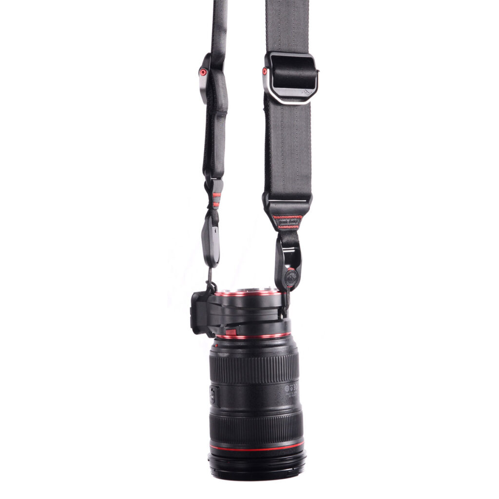 Peak Design Lens Kit - Canon EF, jossa on musta objektiivi punaisilla korostuksilla, on kiinnitetty pystysuoraan leveään, säädettävään mustaan hihnaan, jossa on metalliklipsit ja -kiinnikkeet, valkoista taustaa vasten.