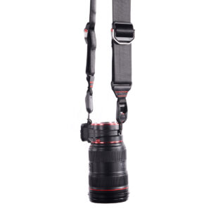 Peak Design Lens Kit - Canon EF, jossa on musta objektiivi punaisilla korostuksilla, on kiinnitetty pystysuoraan leveään, säädettävään mustaan hihnaan, jossa on metalliklipsit ja -kiinnikkeet, valkoista taustaa vasten.