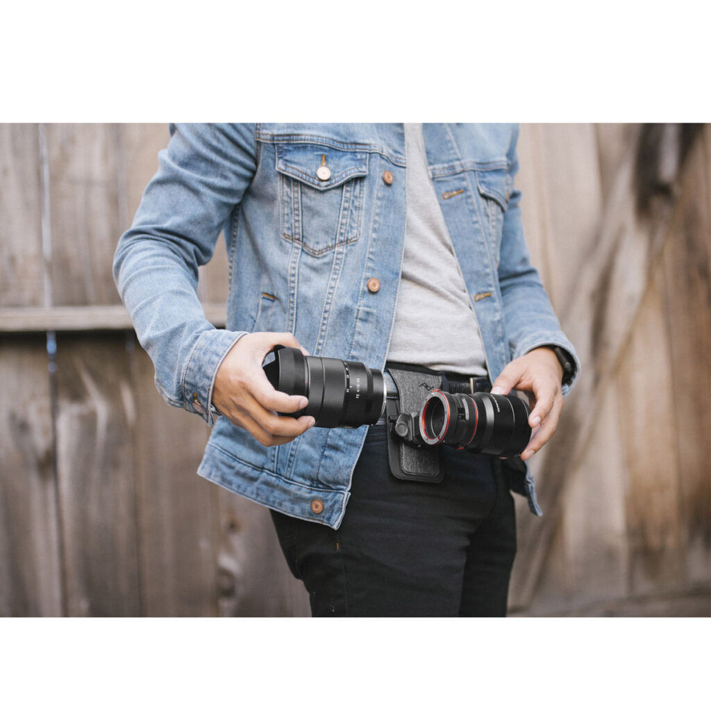 Farkkutakki päällä ja puisen aidan edessä seisova henkilö vaihtaa Canon EF -objektiiveja Peak Design Lens Kit -objektiivisarjaan, jokainen objektiivi kädessä, samalla kun hänellä on vyötärönsä ympärillä kamerakotelovyö.