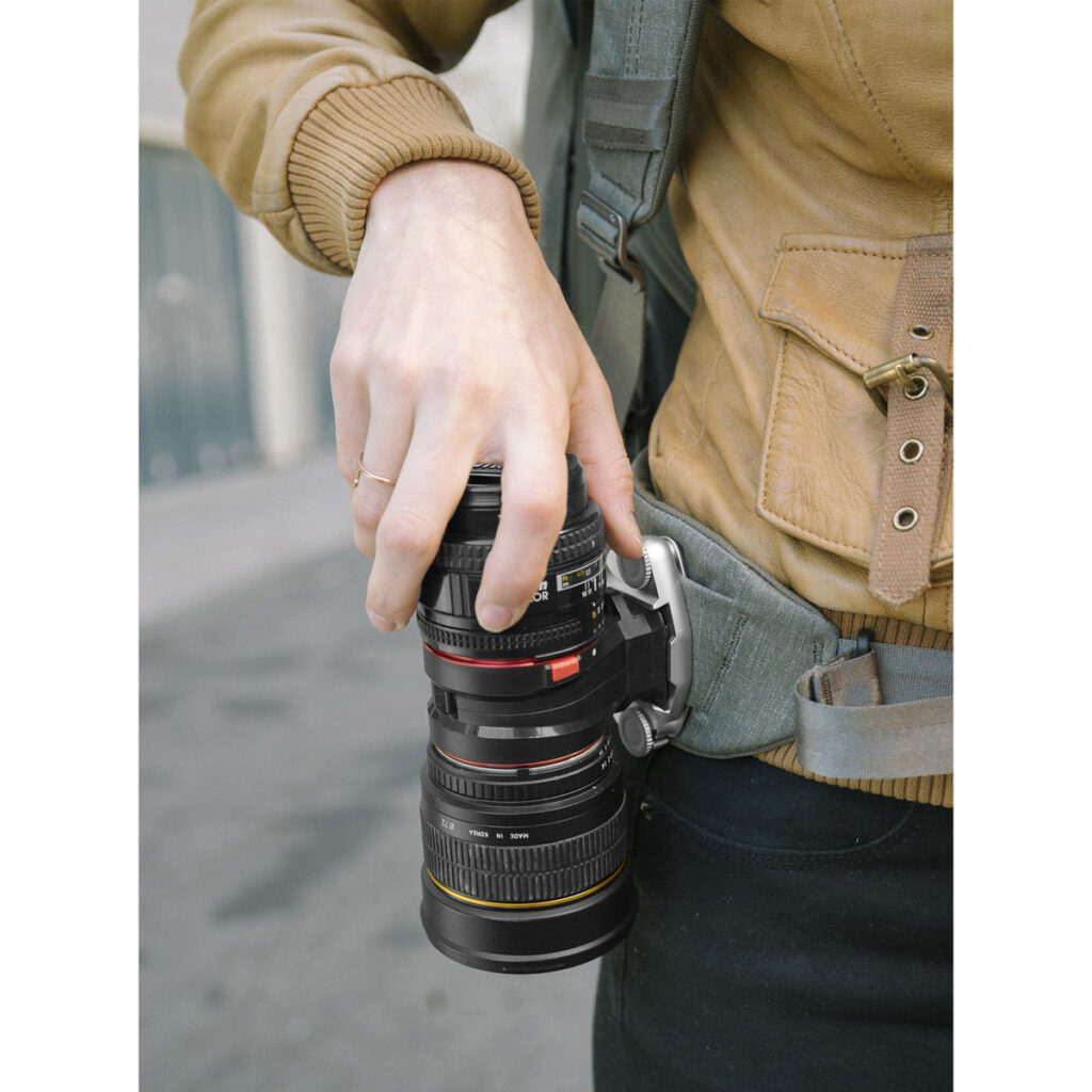 Ulkona kaupungin kadulla ruskeaan takkiin pukeutunut henkilö käyttää Peak Design Lens Kit - Canon EF -objektiivisarjaa kiinnittääkseen kameran objektiivin turvallisesti vyöhönsä siihen tarkoitetulla pidikkeellä.