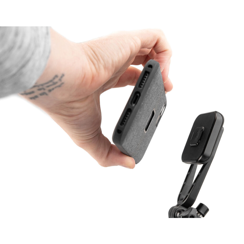 Peak-Design-Mobile-Creator-Kit-5 Käsi pitää älypuhelinta kangaskotelossa lähellä Peak Design Mobile Creator Kit - Black -magneettikiinnikettä jalustalla, jossa näkyvät puhelimen portit ja kaiuttimet.