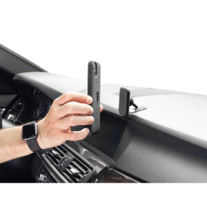 Peak-Design-Mobile-Magnetic-Car-Mount3 Käsi pitää älypuhelinta lähellä Peak Design Mobile VHB-tarra-autotelinettä auton kojelaudalla, valmiina puhelimen kiinnittämiseen. Myös auton sisätilat ja osa henkilön ranteessa olevasta älykellosta näkyvät.