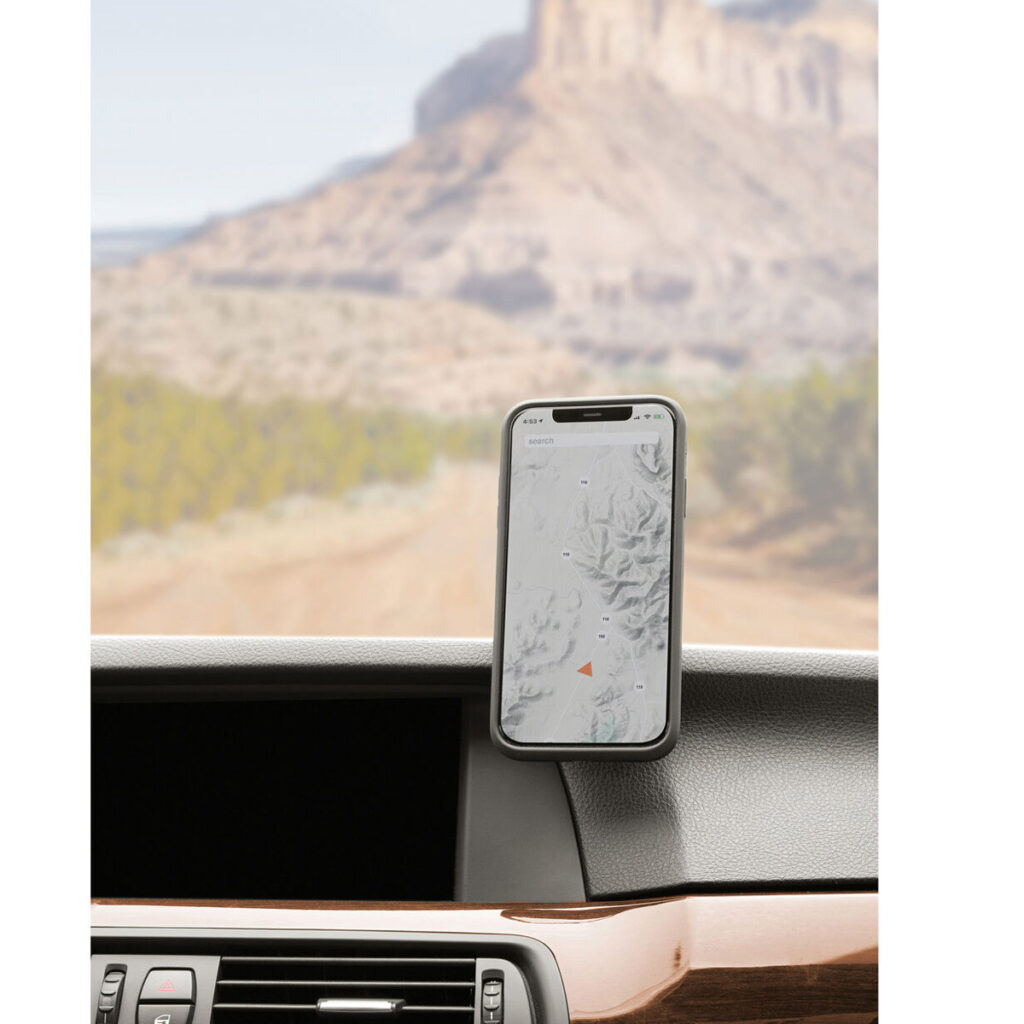 Peak-Design-Mobile-Magnetic-Car-Mount5 Peak Design Mobile VHB-tarra-autotelineessä on älypuhelin, joka näyttää kartan auton kojelaudalla, ja taustalla näkyy tuulilasin läpi sumea kalliomaisema ja hiekkatie.