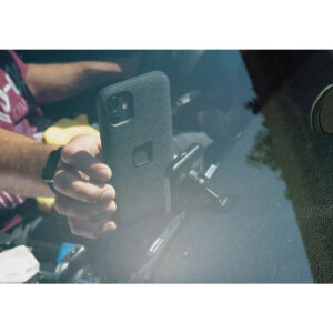 Peak-Design-Mobile-Magnetic-Charging-Car-Mount7 Autossa istuva henkilö kiinnittää mustalla kotelolla varustetun älypuhelimen Peak Design Mobile VHB-tarralatausautotelineeseen kojelaudassa. Tuulilasin läpi otetussa kuvassa näkyy osa kädestä ja heijastukset lasissa.