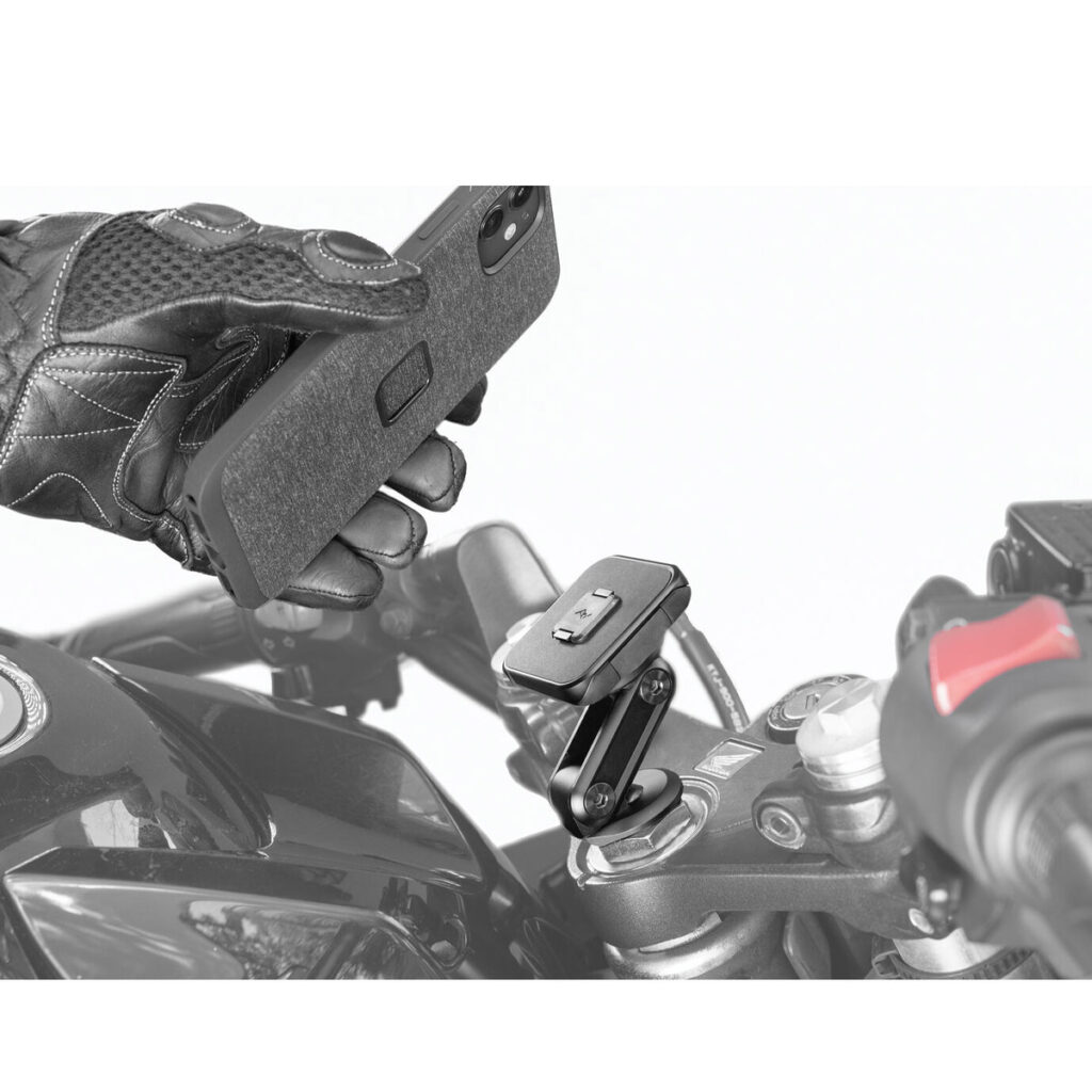 Peak-Design-Mobile-Motorcycle-Stem-Mount-4 Käsi pitää älypuhelinta moottoripyörän ohjaustangon yläpuolella, kun se aiotaan kiinnittää Peak Design Mobile Motorcycle Stem Mount -malliin, ja kiinnike ja pyörän hallintalaitteet ovat selvästi näkyvissä.