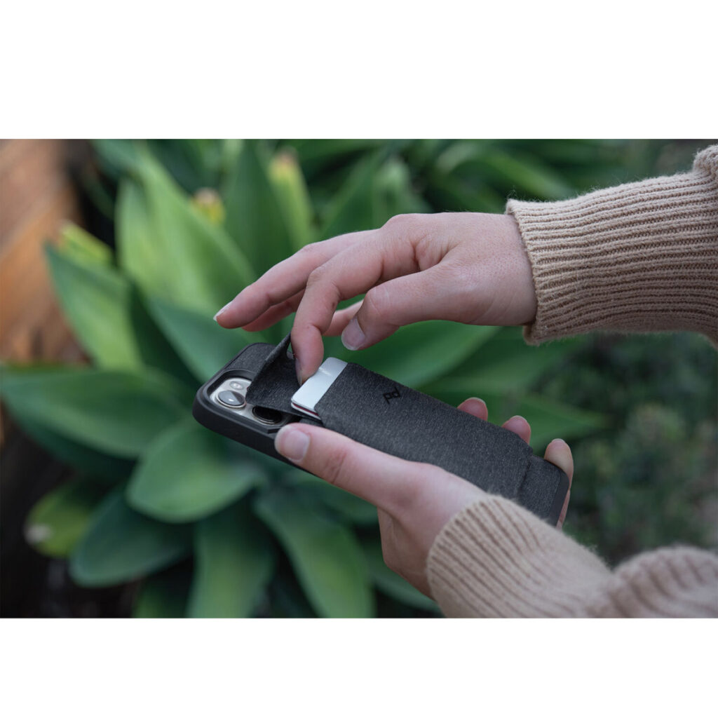 Peak-Design-Mobile-Stand-Wallet-5 Henkilö työntää kortin puhelimeen kiinnitetyn Peak Design Mobile Wallet Stand - Charcoal -lompakkotelineen takataskuun, taustalla vihreitä kasveja.