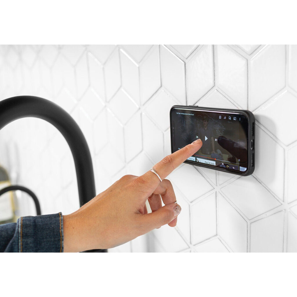 Peak-Design-Mobile-Wall-Mount-Bone-4 Käsi napauttaa älypuhelinta Peak Design Mobile Wall Mount - Bone -seinäkiinnikkeessä, joka on kiinnitetty valkoiseen geometriseen laattaseinään mustan hanan läheisyyteen; puhelin näyttää pysäytetyn videon.