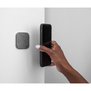 Peak-Design-Mobile-Wall-Mount-Charcoal-2 Henkilö pitää älypuhelinta lähellä Peak Design Mobile Wall Mount - Charcoal -seinäkiinnikettä, joka on pieni, neliönmuotoinen laite, joka on kiinnitetty valkoiselle seinälle.