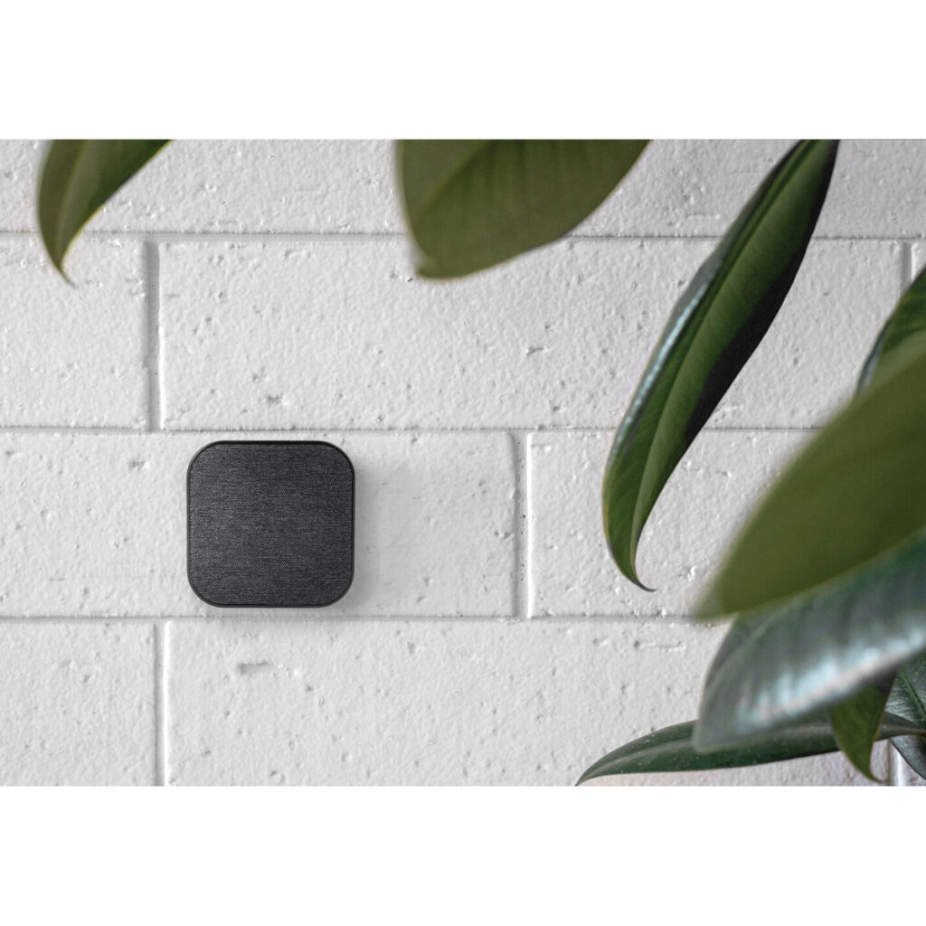 Peak-Design-Mobile-Wall-Mount-Charcoal-3 Peak Design Mobile Wall Mount - Charcoal, pieni musta neliönmuotoinen laite, on asennettu valkoiselle tiiliseinälle, ja suuret vihreät kasvinlehdet kehystävät osittain kuvan oikeaa reunaa.