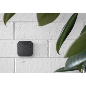 Peak-Design-Mobile-Wall-Mount-Charcoal-3 Peak Design Mobile Wall Mount - Charcoal, pieni musta neliönmuotoinen laite, on asennettu valkoiselle tiiliseinälle, ja suuret vihreät kasvinlehdet kehystävät osittain kuvan oikeaa reunaa.