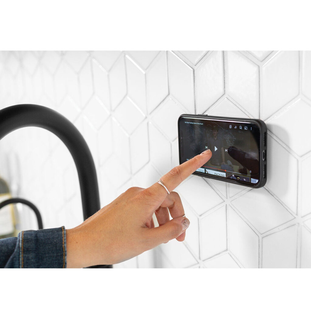 Peak-Design-Mobile-Wall-Mount-Charcoal-5 Käsi, jossa on hopeinen rengas, napauttaa älypuhelinta, joka on kiinnitetty Peak Design Mobile Wall Mount - Charcoal -seinätelineellä valkoiselle kuusikulmaiselle laattaseinälle. Puhelin näyttää videon, ja vasemmalla näkyy osa mustasta keittiöhanasta.