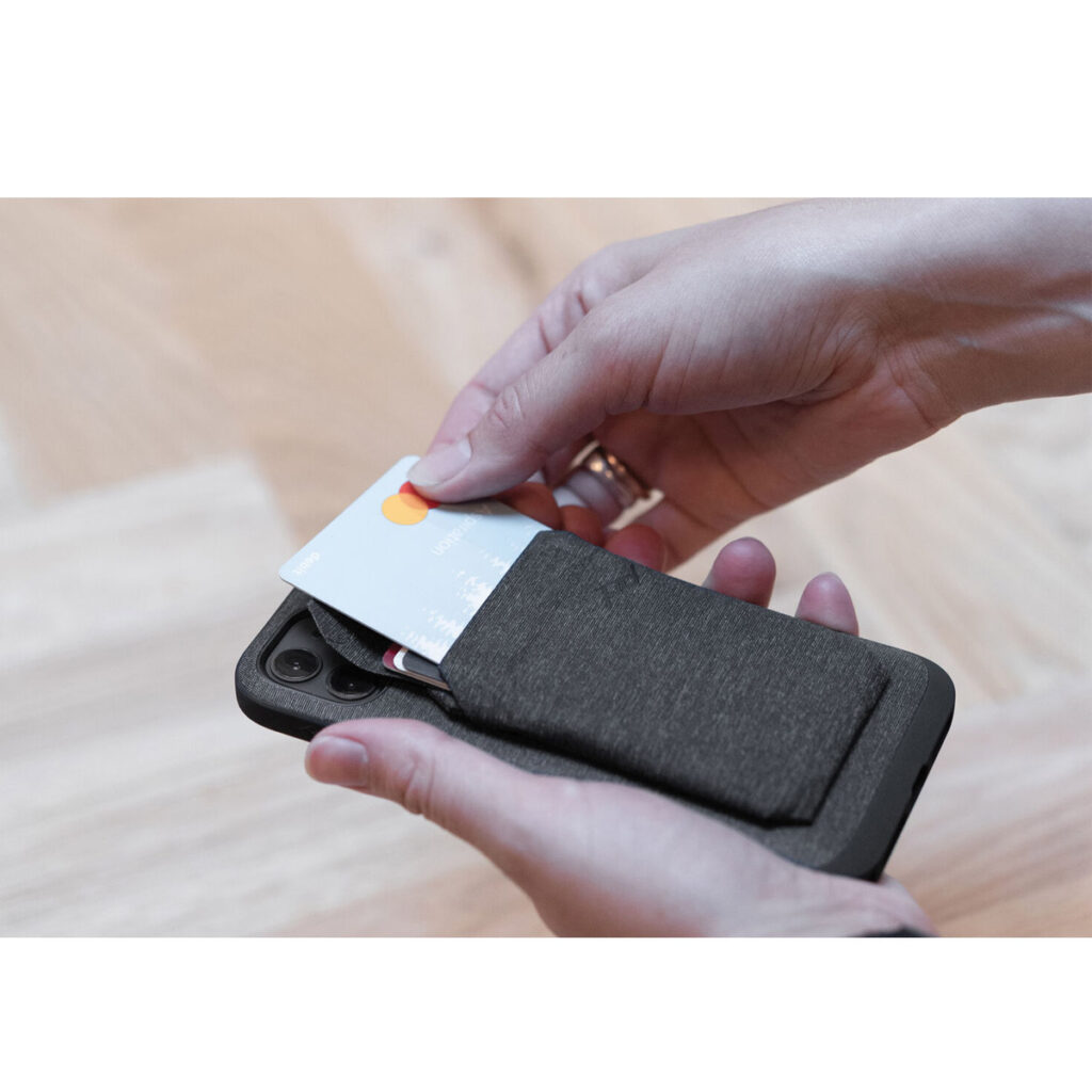 Peak-Design-Mobile-Wallet-Slim-5 Henkilö asettaa luottokortin Peak Design Mobile Wallet Slim - Charcoal -lompakkoon, joka on kiinnitetty mustan älypuhelimen takaosaan, ja taustalla on vaalea puulattia.