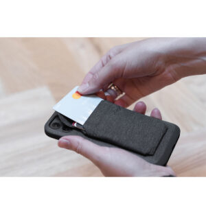 Peak-Design-Mobile-Wallet-Slim-5 Henkilö asettaa luottokortin Peak Design Mobile Wallet Slim - Charcoal -lompakkoon, joka on kiinnitetty mustan älypuhelimen takaosaan, ja taustalla on vaalea puulattia.