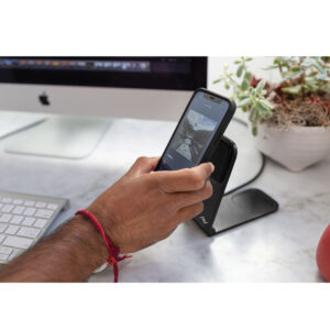 Peak-Design-Mobile-Wireless-Charging-Stand-4 Henkilö pitää älypuhelinta pöydällä olevan Peak Design Mobile Wireless Charging Stand -lataustelineen päällä, taustalla iMac, näppäimistö ja ruukkukasvi.